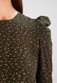 Blusa verde oliva con accenti rotondi a motivo bianco e nero. Presenta maniche a sbuffo e un tessuto liscio e testurizzato.