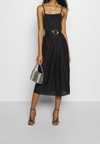 Frau in einem schwarzen, plissierten Midirock und einem schwarzen Spaghetti-Trägertop, die eine kleine Handtasche mit Schlangenhaut-Muster hält und schwarze Sandalen mit Riemchen trägt.
