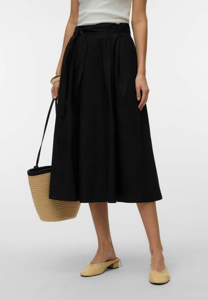 Femme portant une jupe midi noire avec ceinture, chaussures beiges à talons bas, tenant un sac en paille avec des finitions noires.