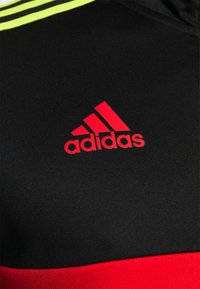 Camiseta deportiva negra con el logo rojo de adidas. Presenta una franja amarilla en el cuello y un detalle rojo en la parte inferior. Textura de tela suave.