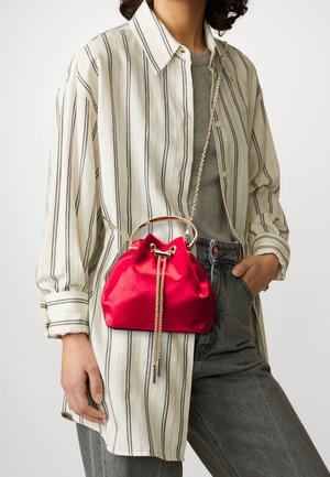 Personne portant une chemise à rayures et un jean gris tenant un petit sac à cordon en satin rouge avec une bandoulière chaîne dorée.