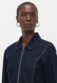 Temno moderna jeans jakna z zadrgo spredaj, špičastim ovratnikom in kontrastnim belim šivanjem. Tkanina je gladka z rahlo teksturo.