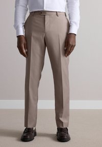 Pantalons habillés beiges avec une taille plate, coupe ajustée, deux poches latérales et une fermeture à un bouton. Associés à des chaussures sombres et une chemise blanche.