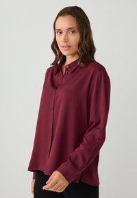Femme aux cheveux bruns ondulés portant une chemise bordeaux à manches longues et boutonnée large, ainsi qu'un pantalon noir, se tenant contre un fond clair.