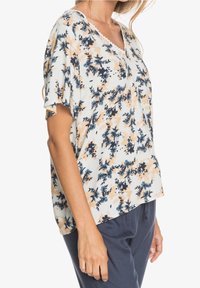 Blusa floral de manga corta en tejido ligero, con flores azules y melocotón sobre un fondo blanco, con escote en forma de V y ribete con puntilla.