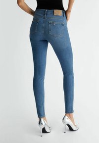 Jeans de mezclilla azul ajustados con una cintura media, bolsillos traseros y un parche de cuero en la parte posterior, combinados con elegantes tacones altos plateados.