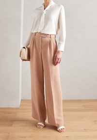 Bílá blůza s uvázaným krkem, béžové kalhoty s vysokým pasem a širokými nohavicemi, malá béžová kabelka a bílé sandály na dřevěné podlaze.