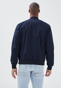 Marinfärgad bomberjacka med elastiska muddar och fåll, med en slät yta och en enkel design, sedd från baksidan.