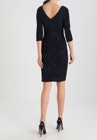 Vestido negro con escote en forma de V en la espalda, mangas tres cuartos y patrón texturizado. Combinado con tacones altos negros, mostrando una silueta ajustada.