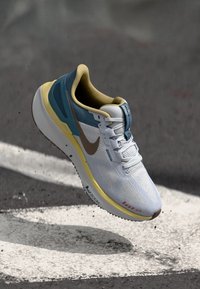 Hvide sportsko med blå og gule accenter, mesh-overdel, struktureret design og brun yttersål. Har Nike-logo og synlig dæmpningsteknologi.