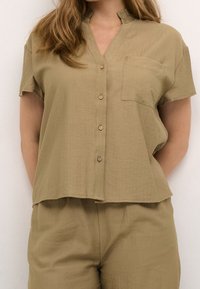 Blusa khaki a maniche corte con scollo a V, chiusura frontale con cinque bottoni e taschino. Tessuto leggero e testurizzato con una vestibilità rilassata.