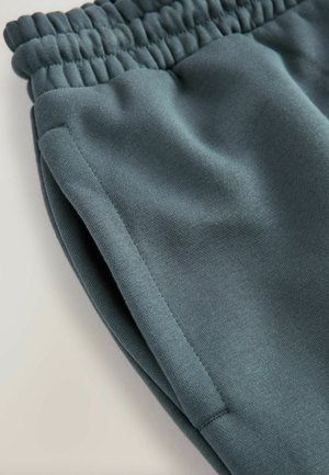 Mīksti, tirkīza krāsas treniņbikses ar elastīgu jostasvietu. Raksturo gluda tekstūra, sānu šuve un savākta jostasvietas detaļa.