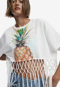 Biely skrátený tričko s farebnou grafikou ananásu, s bielym strapcovým detailom pozdĺž lemovania. Neformálny, uvoľnený strih.