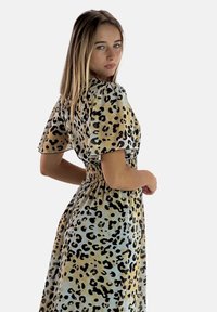 Junge Frau mit langen Haaren, die ein kurzärmeliges, leopardgemustertes Kleid trägt und über ihre Schulter gegen einen neutralen Hintergrund schaut.