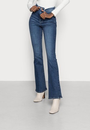 Person, der spænder en knap på blå jeans med høj talje og vidde ben, iført en hvid langærmet top og hvide ankelstøvler med hæl.