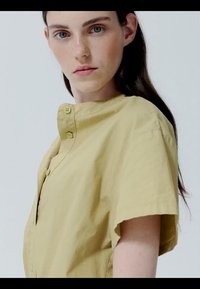 Młoda kobieta o jasnej karnacji i ciemnych włosach, ubrana w jasnożółtą koszulę na krótki rękaw z guzikami, patrząca prosto w obiektyw.