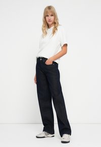 Tamsiai mėlynos plačios džinsinės kelnės su aukštu liemeniu, derinamos su laisvai krentančia balta medvilnine marškinėliais, vilkimos su pilkais sportbačiais.