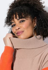 Maglione beige oversize con colletto a coste alto e maniche lunghe a coste arancioni. Il modello ha i capelli ricci e indossa un grande orecchino a cerchio in argento.
