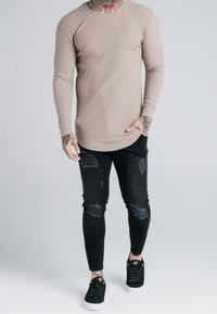 Béžový top s dlouhým rukávem a žebrovaným vzorem, s zakřiveným lemem, spárovaný s černými otrhanými skinny džíny a černými teniskami se zlatými akcenty.