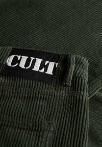 Primer plano de una tela de pana verde que presenta una etiqueta negra con la palabra "CULT" en texto blanco, mostrando la textura de las acanaladuras y los detalles de la costura.