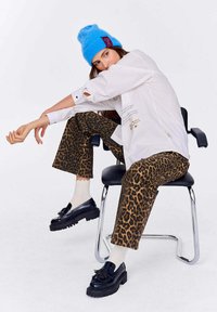 Spodnie w panterkę, biała oversize'owa koszula z wycięciami, niebieska czapka beanie i czarne buty na platformie. Siedząca na metalowym krześle.