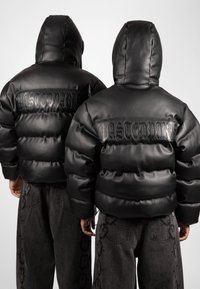Μαύρα πούλεν μπουφάν με γυαλιστερή επιφάνεια, που διαθέτουν το λογότυπο "WASTEDPARIS" στο πίσω μέρος, συνδυασμένα με γκρι τζιν με σχέδια αλυσίδας.