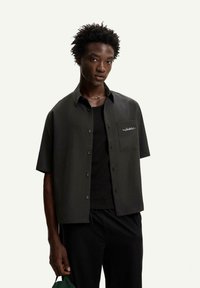 Camicia a maniche corte verde scuro con bottoni, dotata di una tasca sul petto, con logo ricamato bianco e tessuto dalla texture liscia.