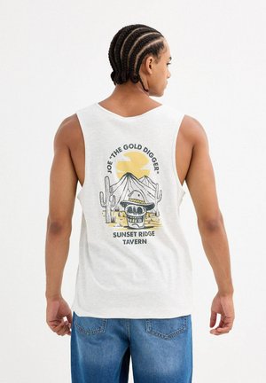 Hombre con cabello trenzado que lleva una camiseta sin mangas blanca con una calavera con sombrero, cactus y montañas bajo un sol amarillo y texto en la parte trasera.