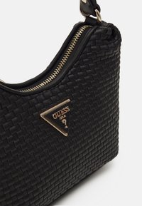 Sac à épaule Guess en cuir tressé noir avec plaque logo triangulaire dorée et fermeture éclair dorée sur le dessus.