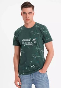 Grøn bomuld T-shirt med rund hals, prydet med hvid tekst: "DIN ENESTE BEGRÆNSNING ER DIN HJERNE," accentueret med en splattermønster.