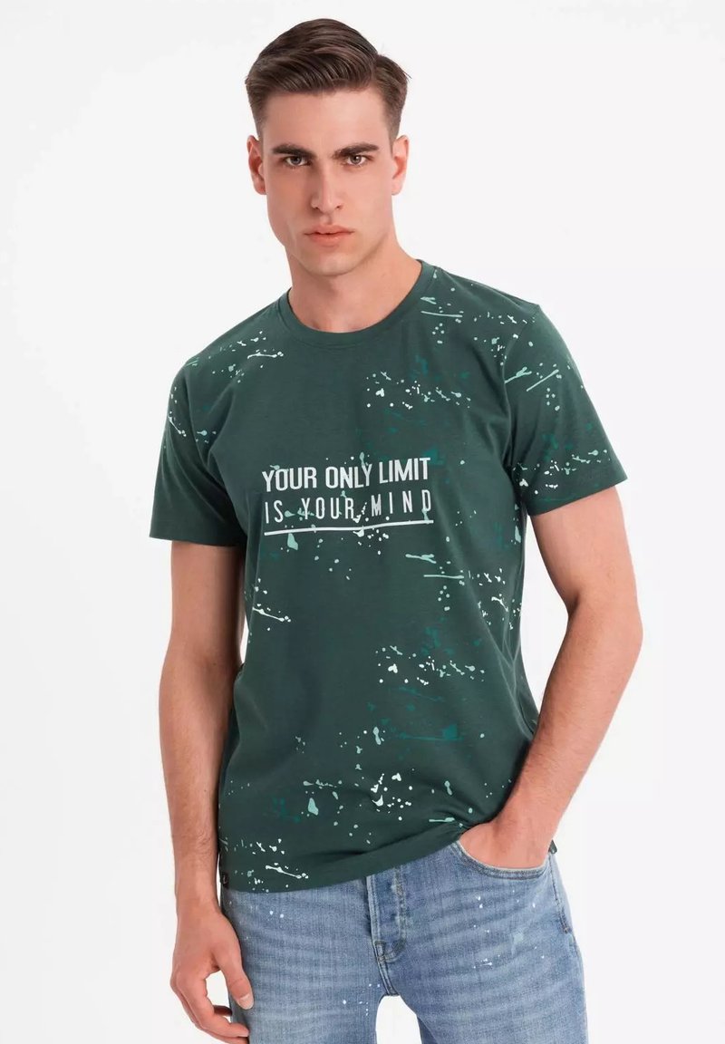 Grøn bomuld T-shirt med rund hals, prydet med hvid tekst: "DIN ENESTE BEGRÆNSNING ER DIN HJERNE," accentueret med en splattermønster.