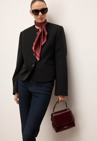 Črn blazer z visokim ovratnikom in enim gumbom, združen z temnimi kavbojkami in rdečim prugastim šalom. Drži bordo torbico z zlatimi dodatki.