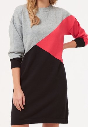 Kvinde iført en farveblokket sweaterkjole med grå, rød og sort diagonale sektioner, lange ærmer og rund halsudskæring.