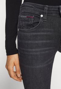 Primer plano de la mano de una persona descansando cerca del bolsillo de unos jeans de mezclilla gris oscuro con un pequeño detalle en rojo, blanco y azul en el bolsillo para monedas.
