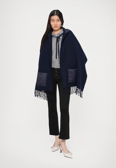 Poncho bleumarin cu margine franjurată, dotat cu buzunare punctate bleumarin. Asortat cu un top gri și pantaloni flared negri. Pantofii albi cu toc completează ținuta.