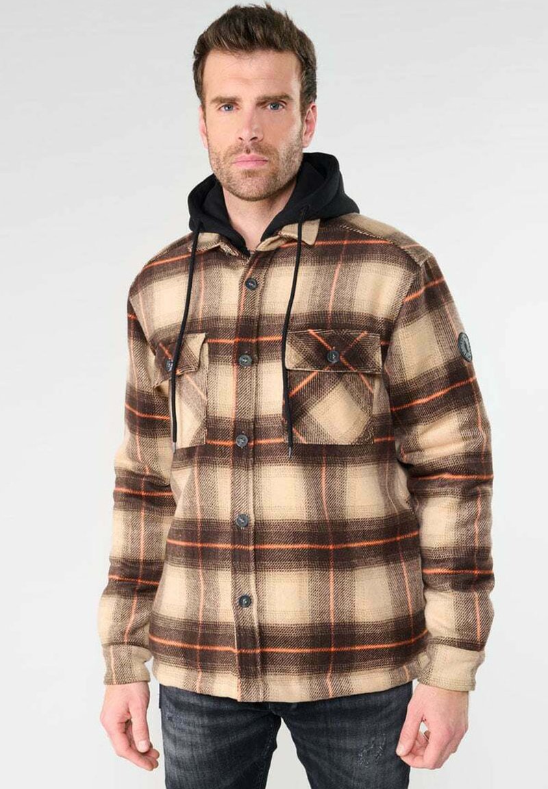 Veste en flanelle à carreaux dans des tons de beige, marron et orange avec une capuche noire. Comprend deux poches poitrine et un devant à boutons. Texture douce.