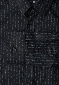 Černá košile s modrým geometrickým vzorem a svislými pruhy. Zapínání na knoflíky a detail kapsy, hladká textura.