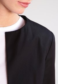 Blazer noir au design épuré et minimaliste, doté d'un col arrondi et d'une texture lisse, porté sur une chemise blanche.