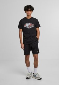 Černé tričko s grafikou "NEPOCTIVÉ BOXOVÁNÍ", spárované s černými šortkami. Model má na sobě sluneční brýle, bílé ponožky a barevné sportovní boty.