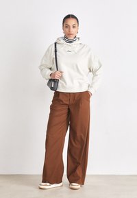 Hanorac crem cu șnururi și logo, pulover cu guler înalt în dungi, pantaloni maro largi, geantă crossbody neagră și adidași albi cu accente maro.