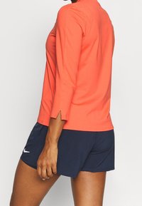 Person in einem korallenfarbenen Langarmshirt mit seitlichen Schlitzen und marineblauen Nike-Sportshorts, der vor einem schlichten Hintergrund steht.