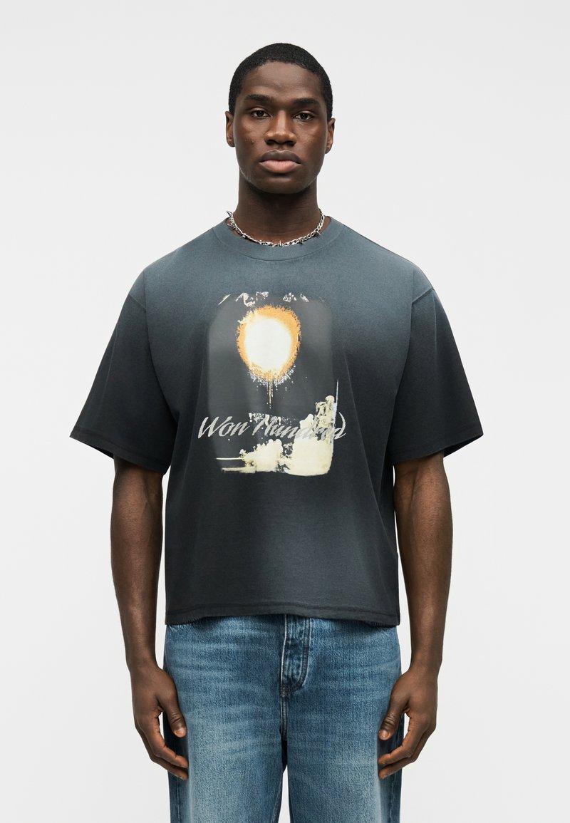 Άνδρας που φοράει σκούρο, φαρδύ T-shirt με γραφικό ήλιο και την επιγραφή «Won Hundred», συνδυασμένο με μπλε τζιν και ασημένιο αλυσίδα λαιμού.