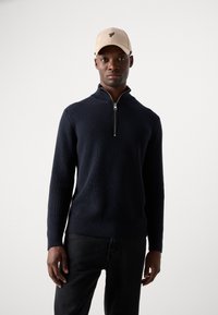 Mørkeblå ribstrikket sweater med høj krave og lynlås, parret med en beige cap med et grønt logo på fronten.
