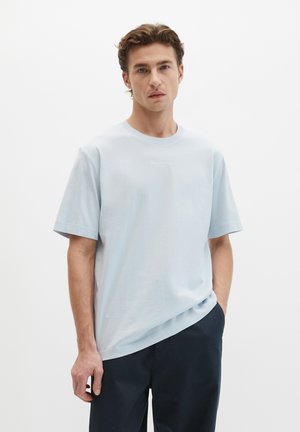 Homme portant un T-shirt bleu clair surdimensionné et un pantalon foncé, debout avec une main dans la poche sur un fond blanc uni.