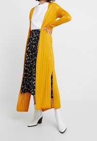 Dlouhý žlutý žebrovaný cardigan s otevřeným předním dílem a bočními rozparky, nošený přes tmavou květovanou sukní s bílým tričkem a bílými botami na podpatku.