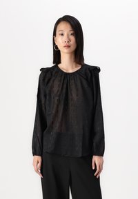 Μαύρη μπλούζα με υφασμάτινη υφή από μικρούς ορθογώνιους σχηματισμούς, με φουσκωτά μανίκια και στρογγυλή λαιμόκοψη, συνδυασμένη με μαύρο παντελόνι.