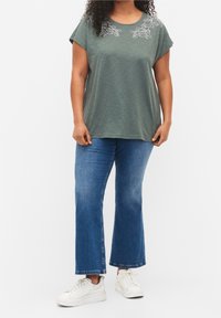 Kvinna iförd en grön blommönstrad kortärmad topp, blå avklippta flarejeans och vita sneakers som står mot en vit bakgrund.