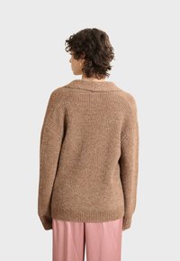 Bruine gebreide trui met een ontspannen pasvorm, geribbelde textuur en brede kraag. Draagt over roze broek, casual achteraanzicht.