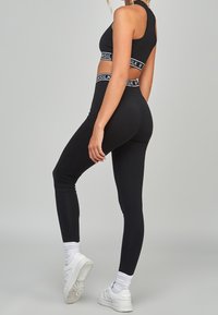 Modell som bär en svart sportoutfit med ett märkesbälte, poserar med en neutral bakgrund och visar upp passformen och stilen.