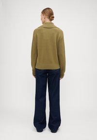 Suéter tricotado verde-oliva com gola alta, punhos e bainha canelados, combinado com jeans de perna larga azul-escuro.
